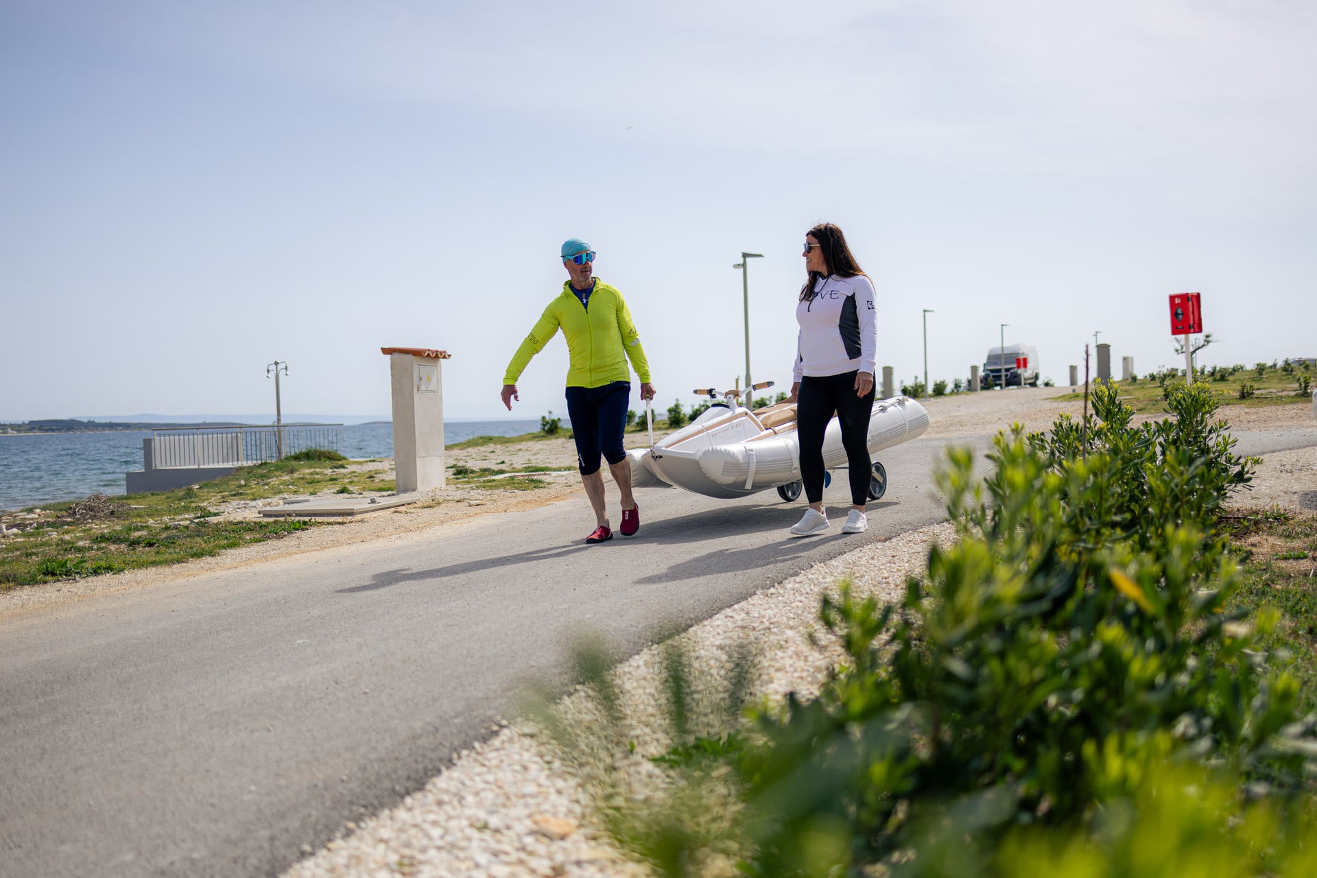 Two people walking on a path by the sea with a vehicle in the background.
